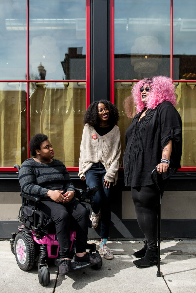 Three Black and disabled folx (a non-binary person in a power wheelchair, a femme leaning against a wall, and a non-binary person standing with a cane) engaged in converation. All three are outdoors and in front of a building with two large windows.