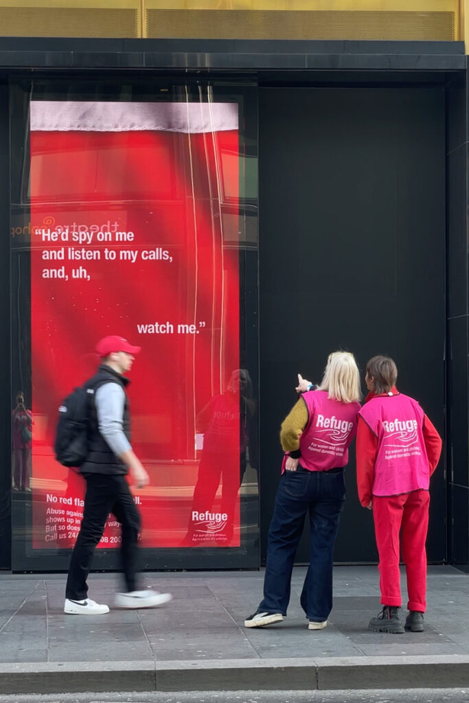 Refuge staff stand with the back to the camera pointing at a long tall Refuge advert as a passer by walks past. The advert reads "He'd spy on me and listen to my calls, and, uh, watch me."