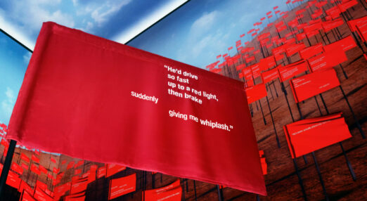 Red flag on a pole reading "He'd drive so fast up to a red light, then brake suddenly giving me whiplash". Behind it is a screen filled with red flags