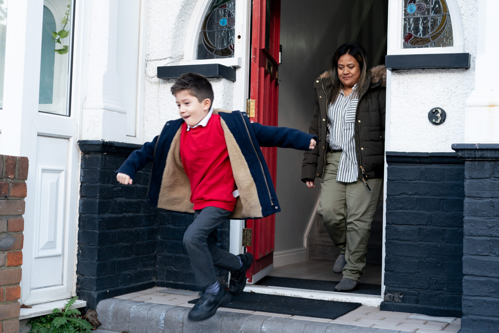 Young boy in red school uniform and a coat runs excitedly out of the front door, followed by his mother who is smiling at him.