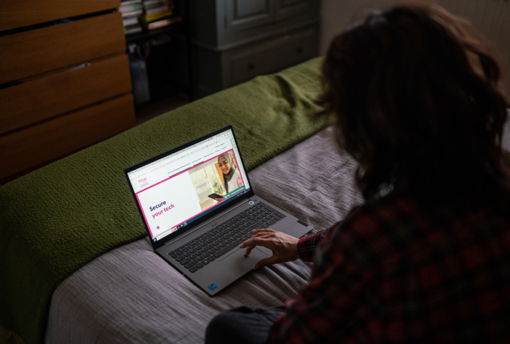 Woman sat on bed with laptop. Webpage shows Refuge's Tech Safety website.